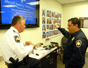 Sheriff James Pohlman and Maj. Chad Clark discuss the drug round-up before addressing assembled officers who will participate. In background are photos of the suspects to be arrested. 