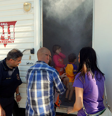 Smoke pours from the open door of the Fire Department mobile safety house as a family enters as Fire Dept. Capt. Rory Miller stands at left. The house is used to teach children how to drop and roll during a fire to avoid smoke inhalation.